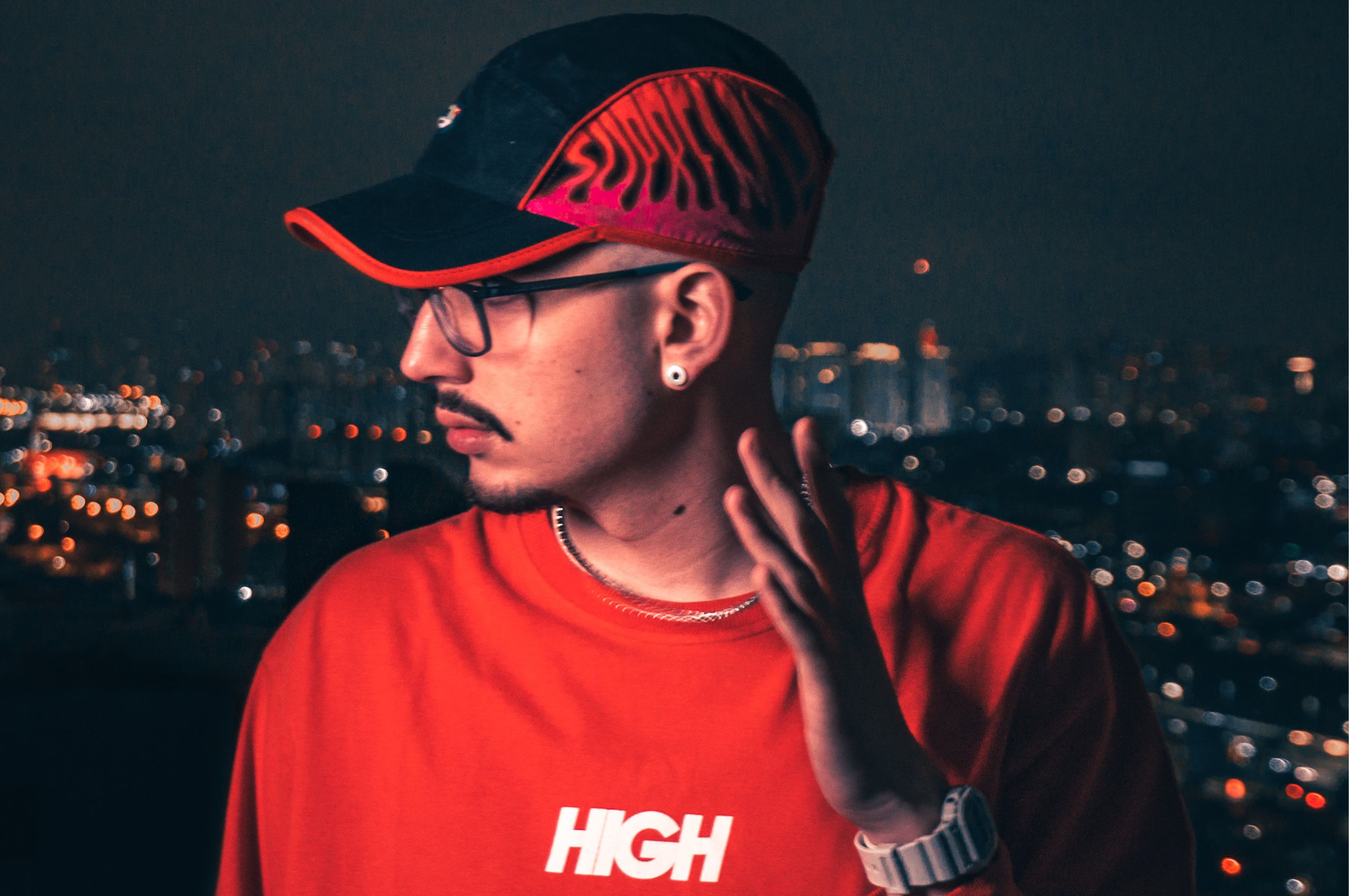Cantor em retrato noturno de perfil, de camiseta vermelha, com as luzes da cidade desfocadas atrás.