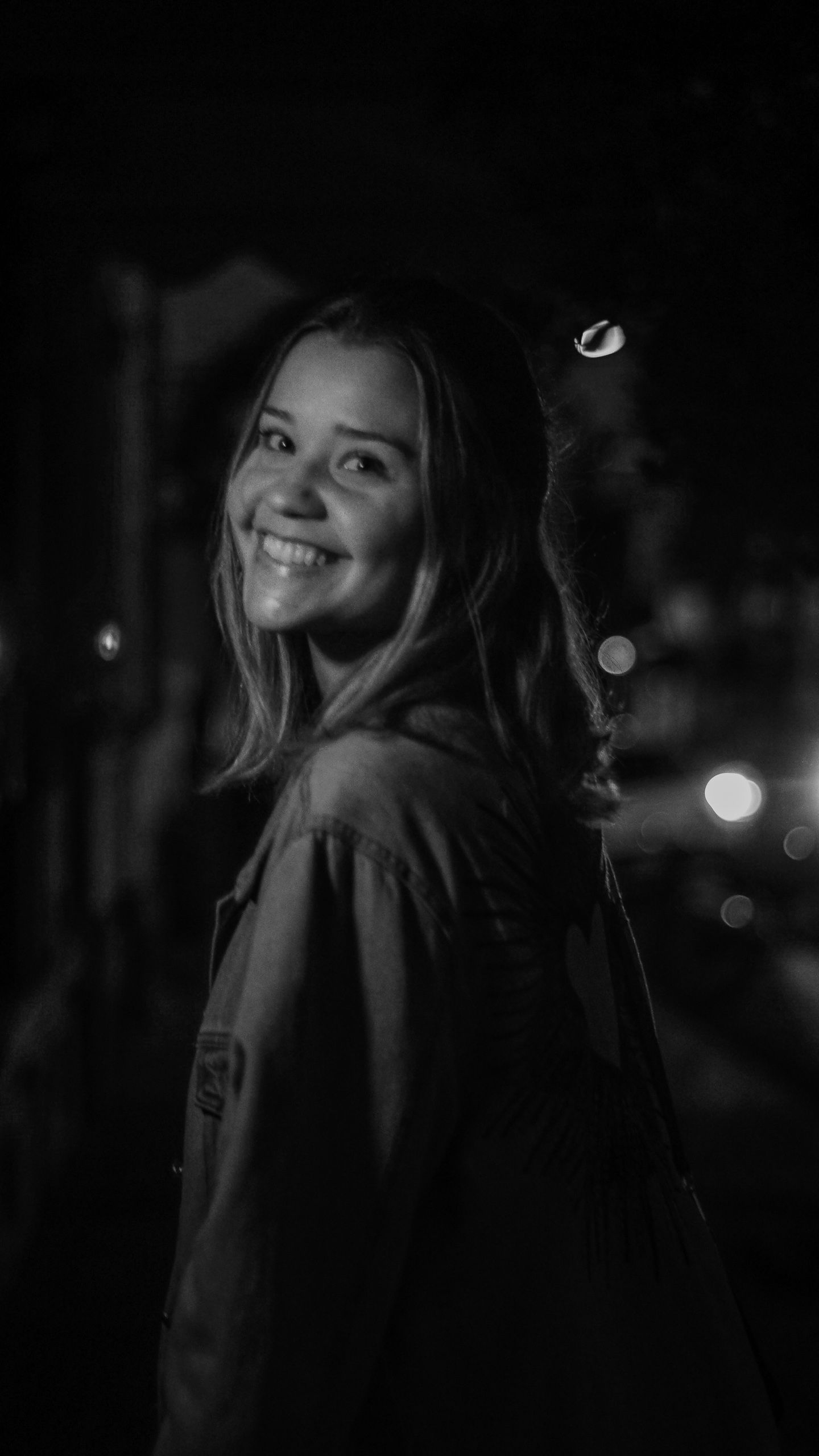 Mulher sorrindo por cima do ombro à noite, em preto e branco com luzes desfocadas.