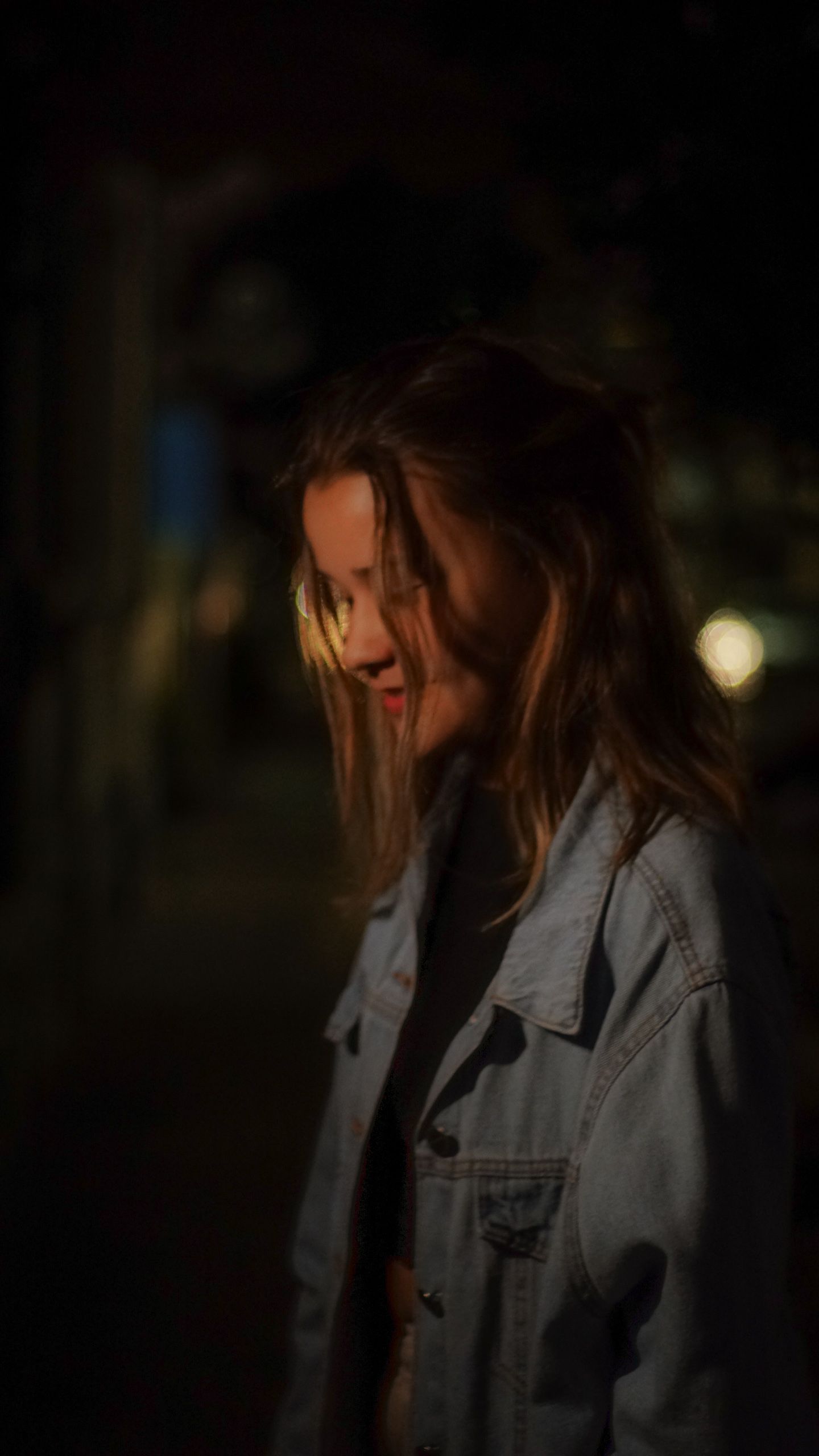 Mulher de jaqueta jeans olhando para baixo à noite, com luz quente no cabelo.