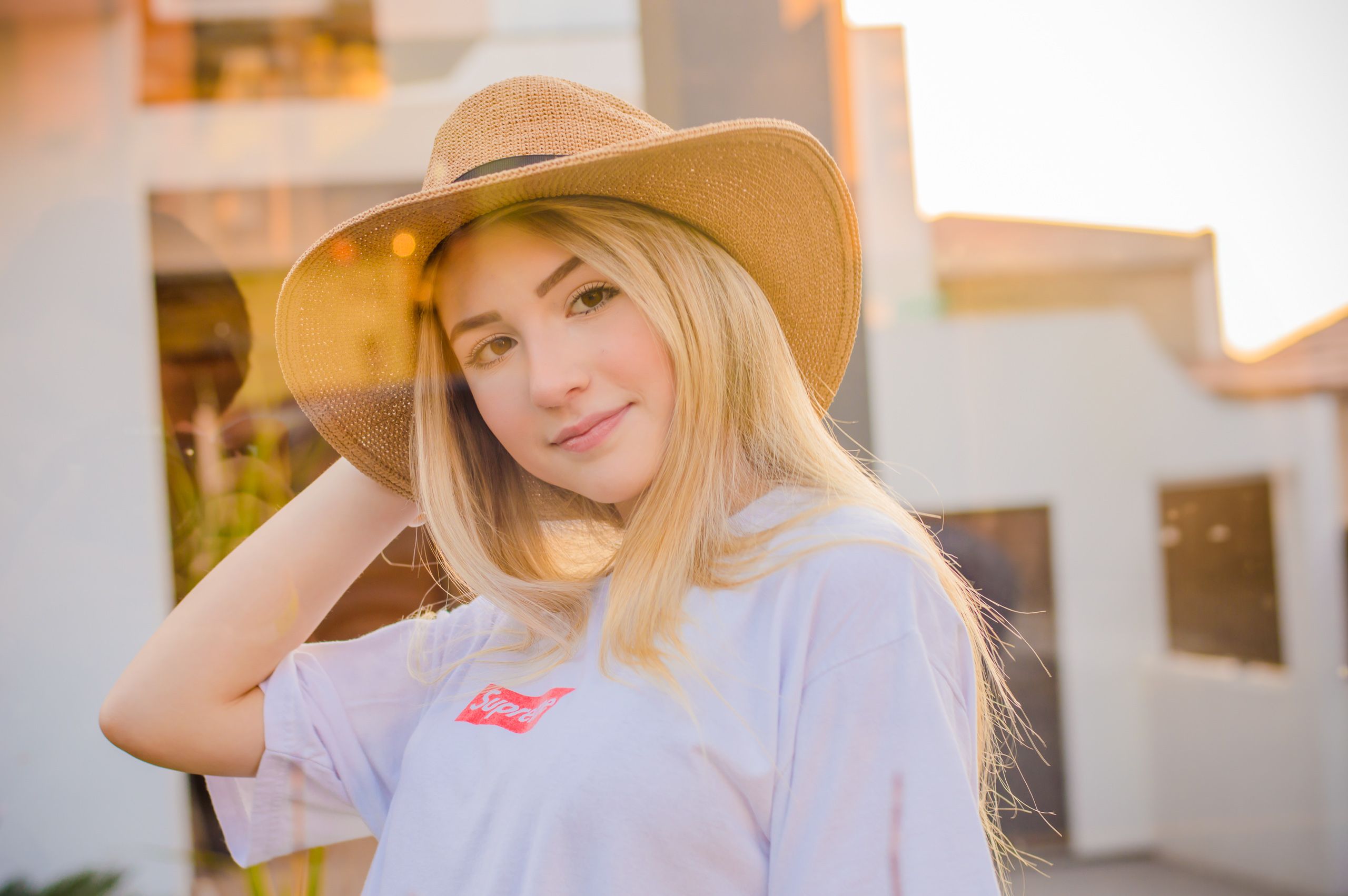 Jovem de cabelo loiro e longo com a mão na aba de um chapéu de palha de abas largas, de camiseta lilás-clara com estampa vermelha, ao ar livre sob luz quente de fim de tarde, com uma construção clara desfocada ao fundo.