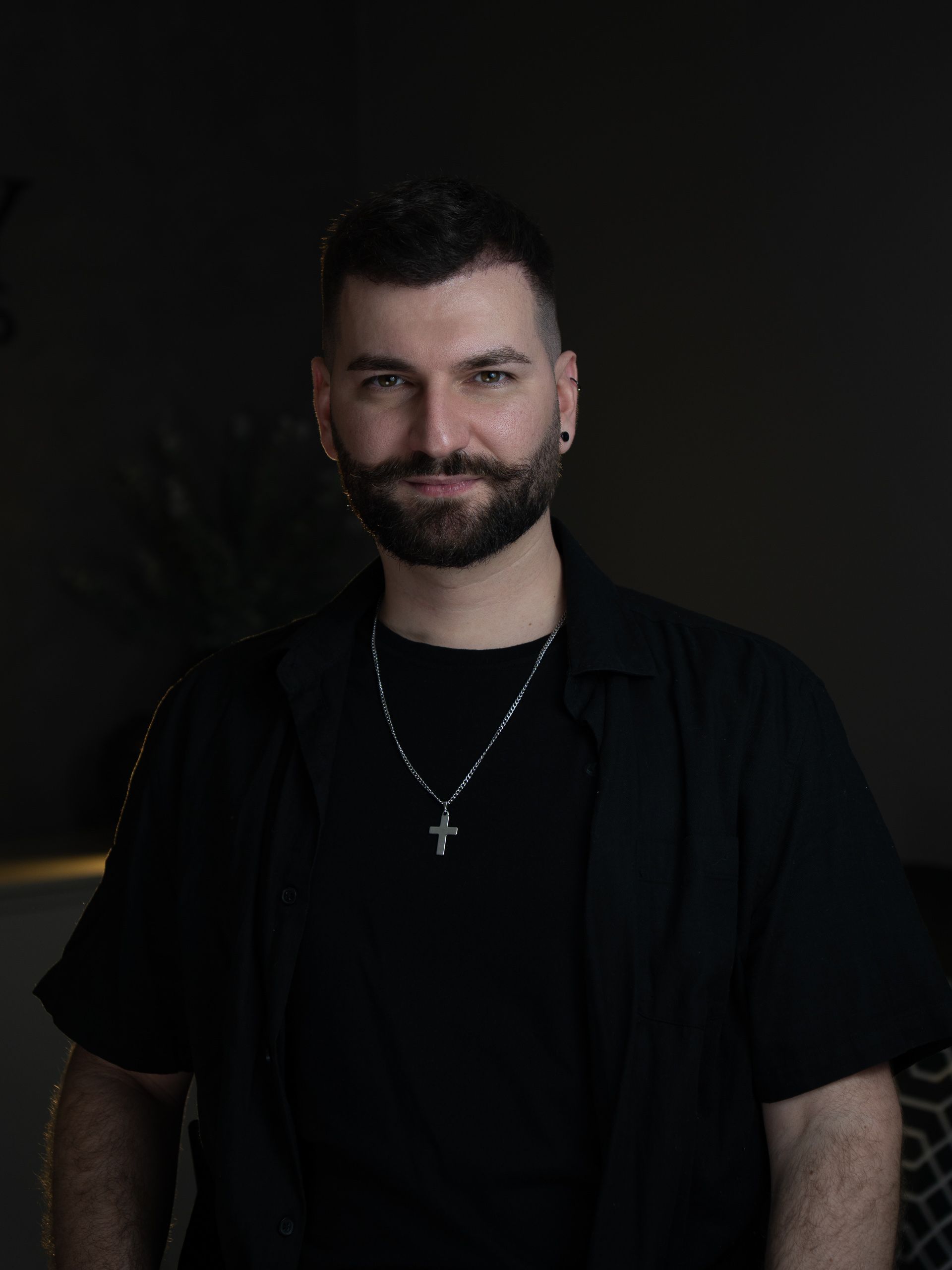 Homem de barba, camisa preta e corrente, em fundo escuro com pouca luz.