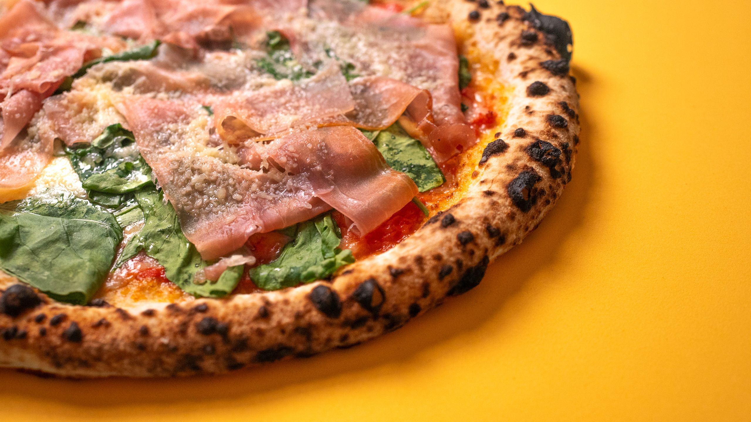 Pizza com fatias de presunto cru e folhas verdes sobre fundo amarelo, em plano fechado.