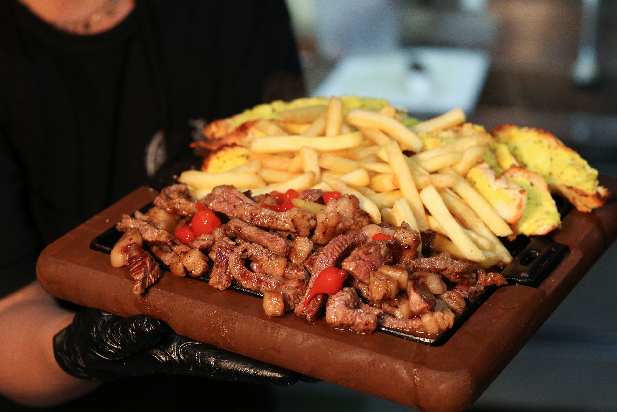 Mão com luva preta segurando tábua com tiras de carne, batatas fritas e pimentas.
