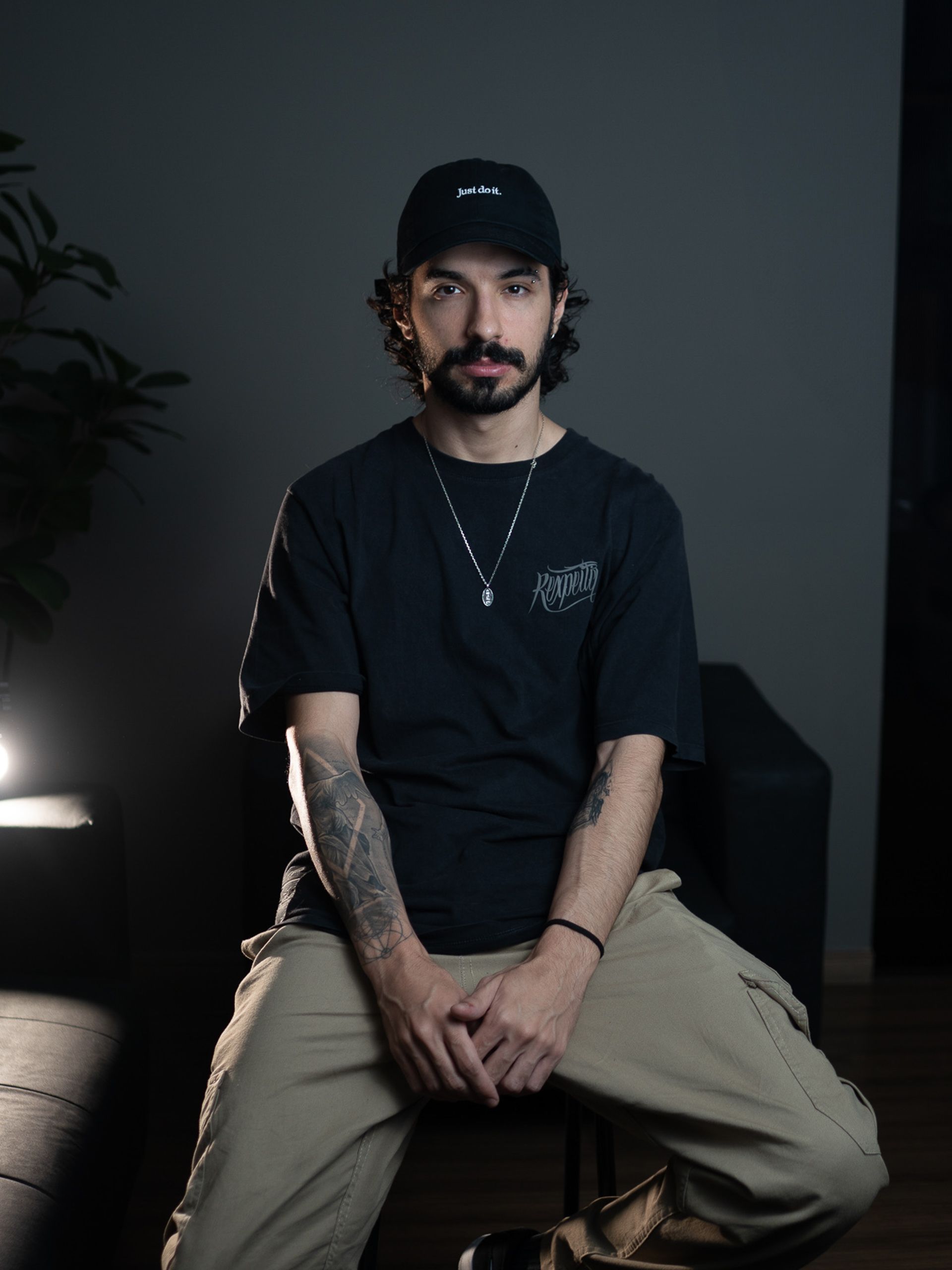Sentado em uma cadeira, jovem tatuado aparece em ambiente de luz baixa, com planta e luminária ao fundo.