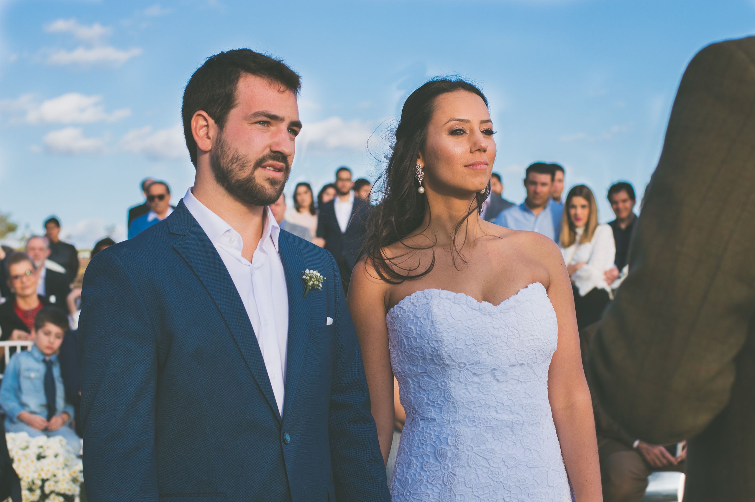 Noivo de terno azul e noiva de vestido branco em cerimônia ao ar livre, com convidados atrás.
