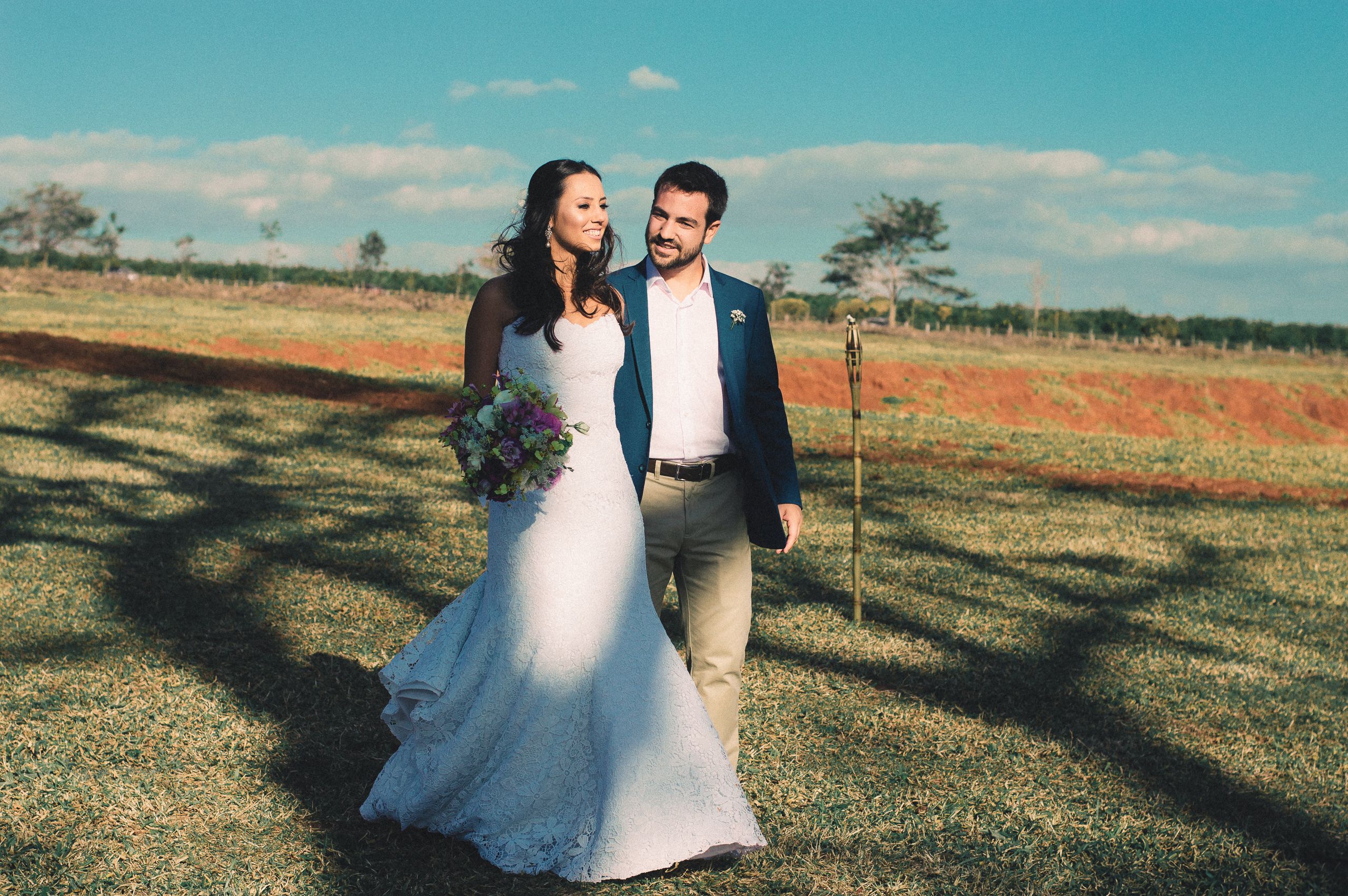 Noiva de vestido longo e homem de blazer azul caminhando por um gramado sob céu azul.