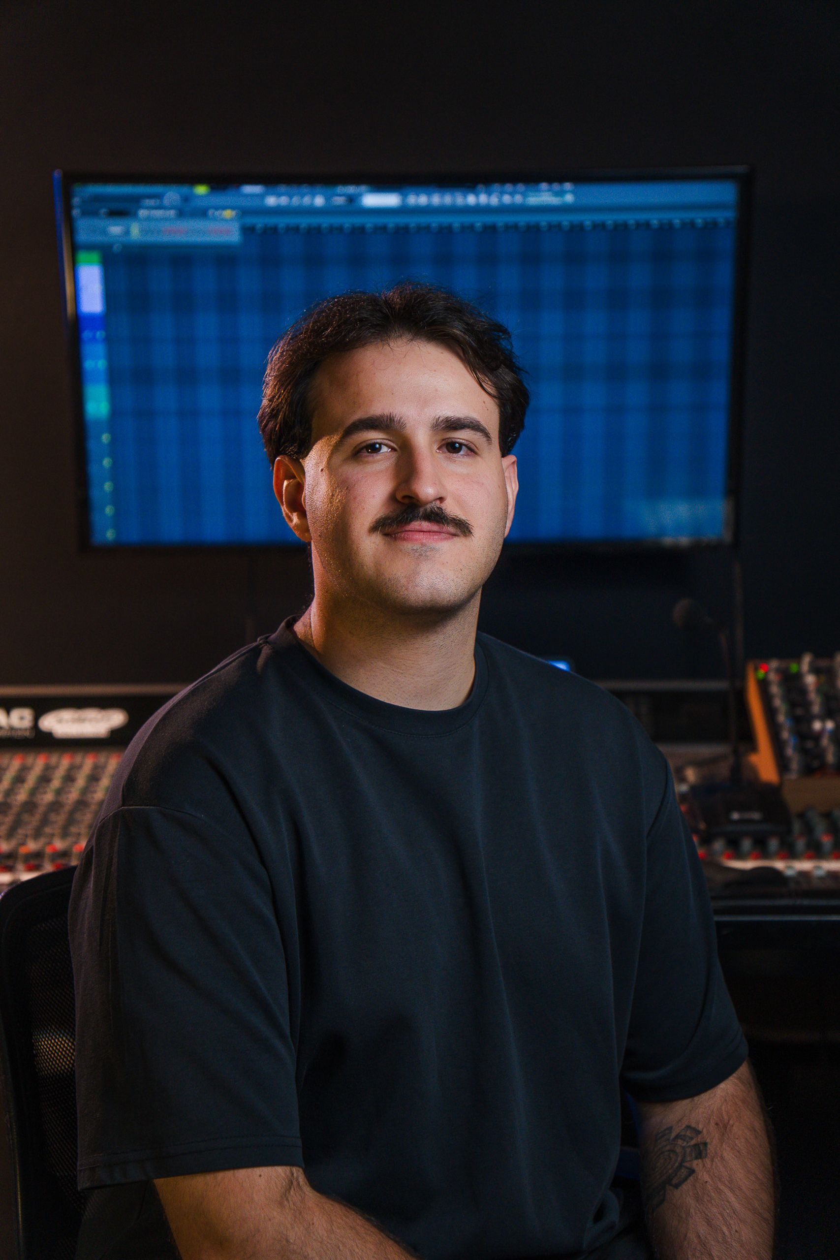 Retrato profissional de produtor musical sentado no estúdio, com tela de edição de áudio atrás.