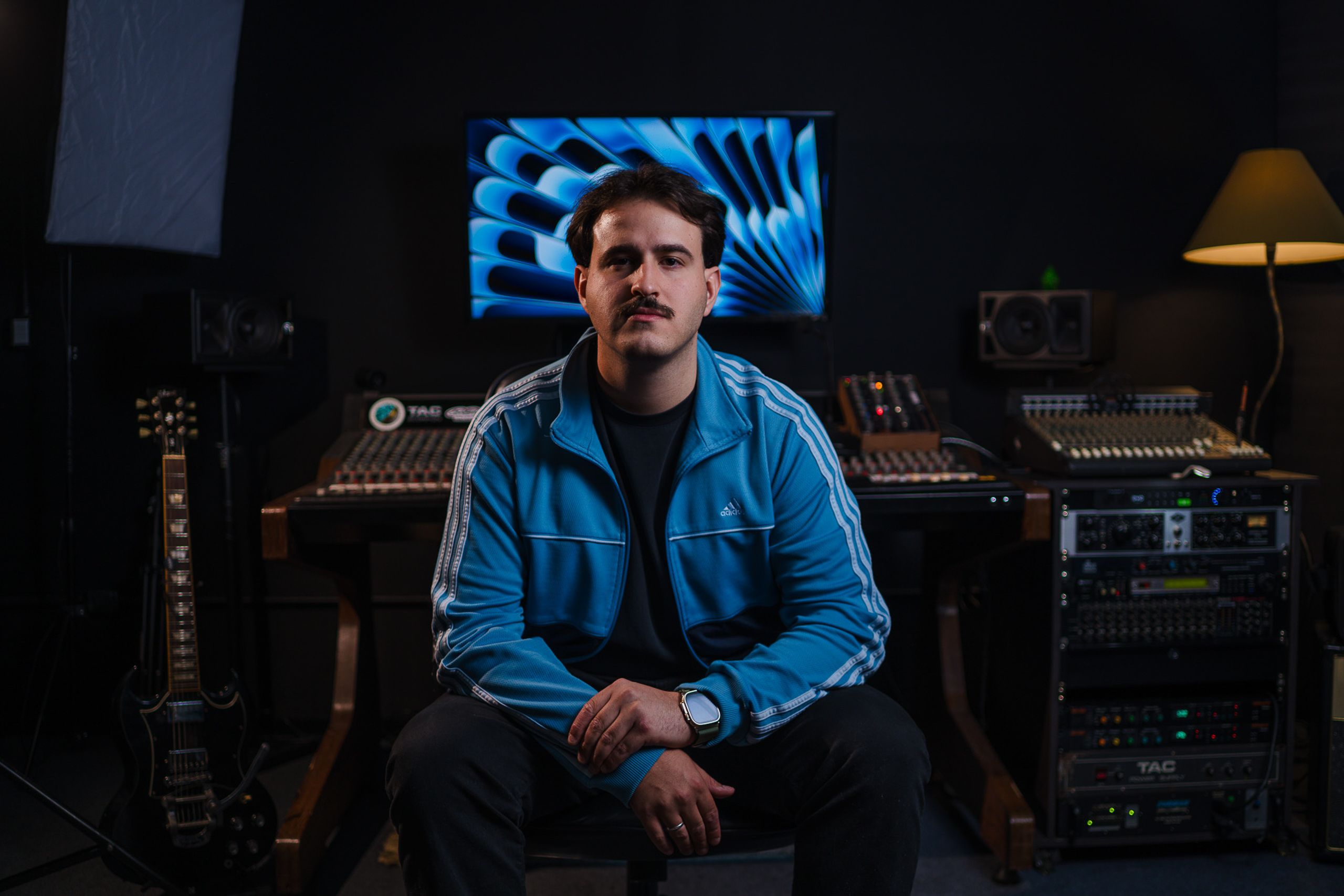 Produtor musical de jaqueta azul sentado no estúdio, com monitor iluminado e guitarra ao lado.