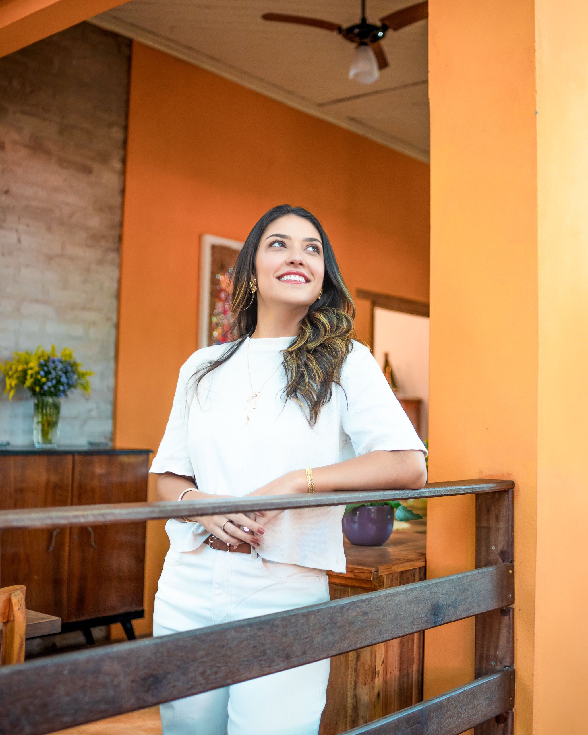 Mulher de branco apoiada em corrimão de madeira, em ambiente de paredes alaranjadas.