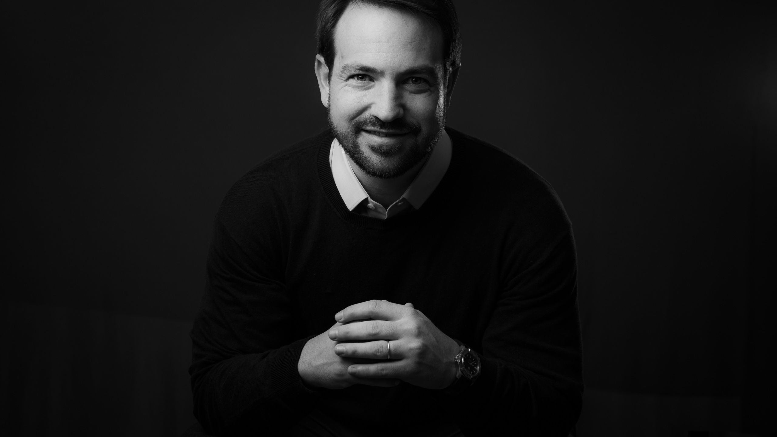 Retrato em preto e branco de homem de suéter e camisa, com as mãos entrelaçadas.