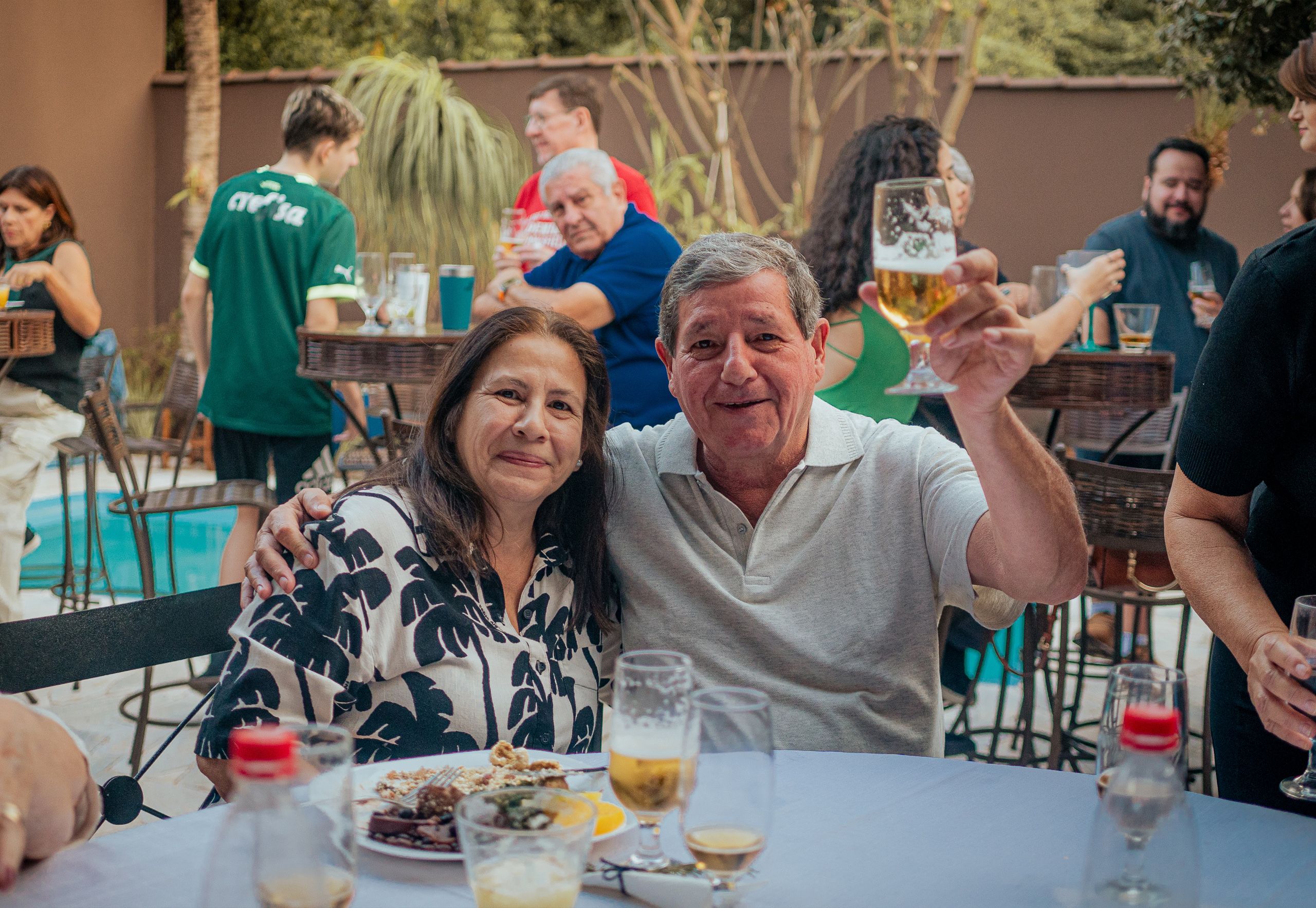 Homem erguendo um copo de cerveja ao lado de mulher sentada, em festa ao ar livre.