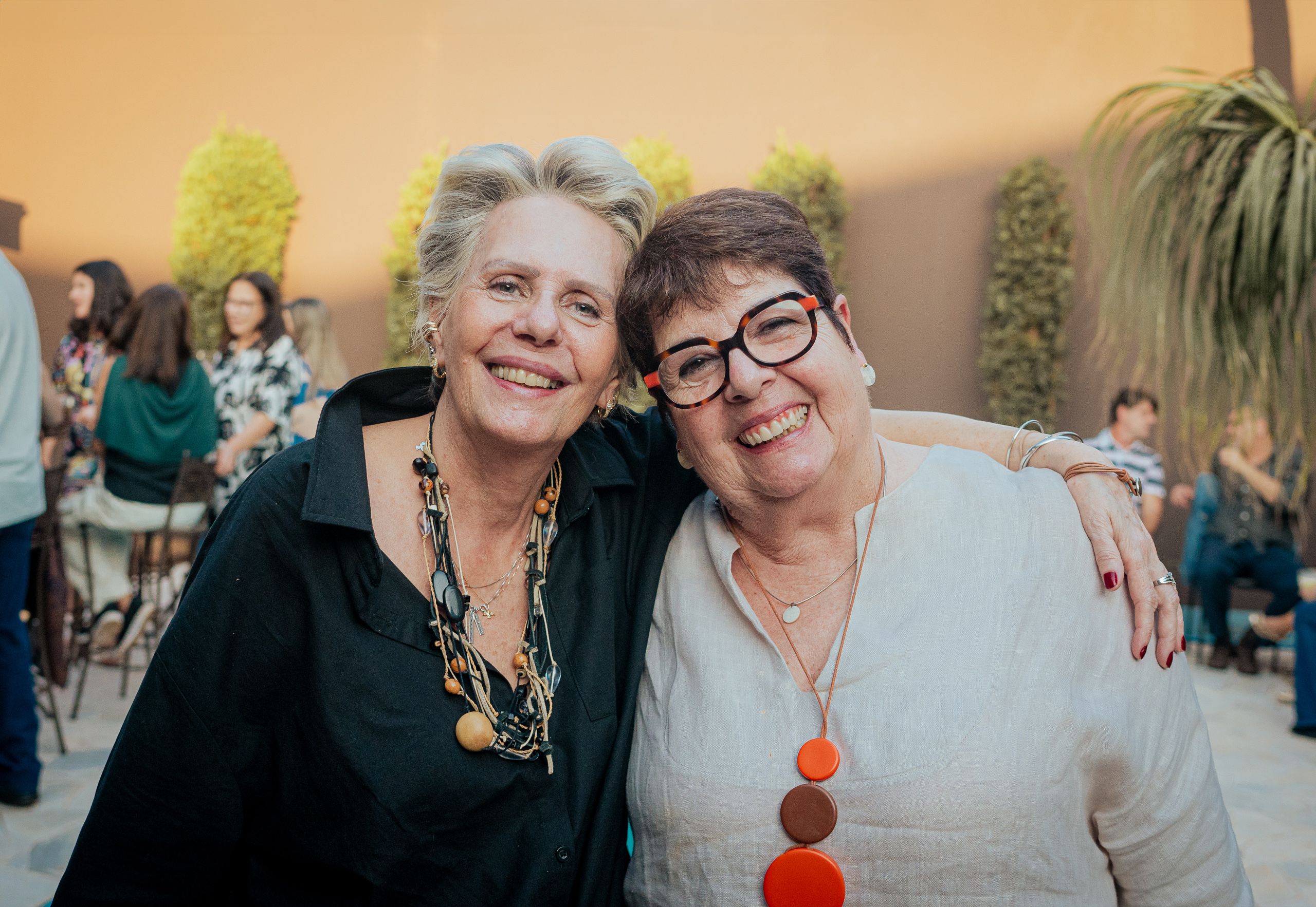 Duas mulheres abraçadas e sorrindo, com convidados desfocados ao fundo em luz de fim de tarde.