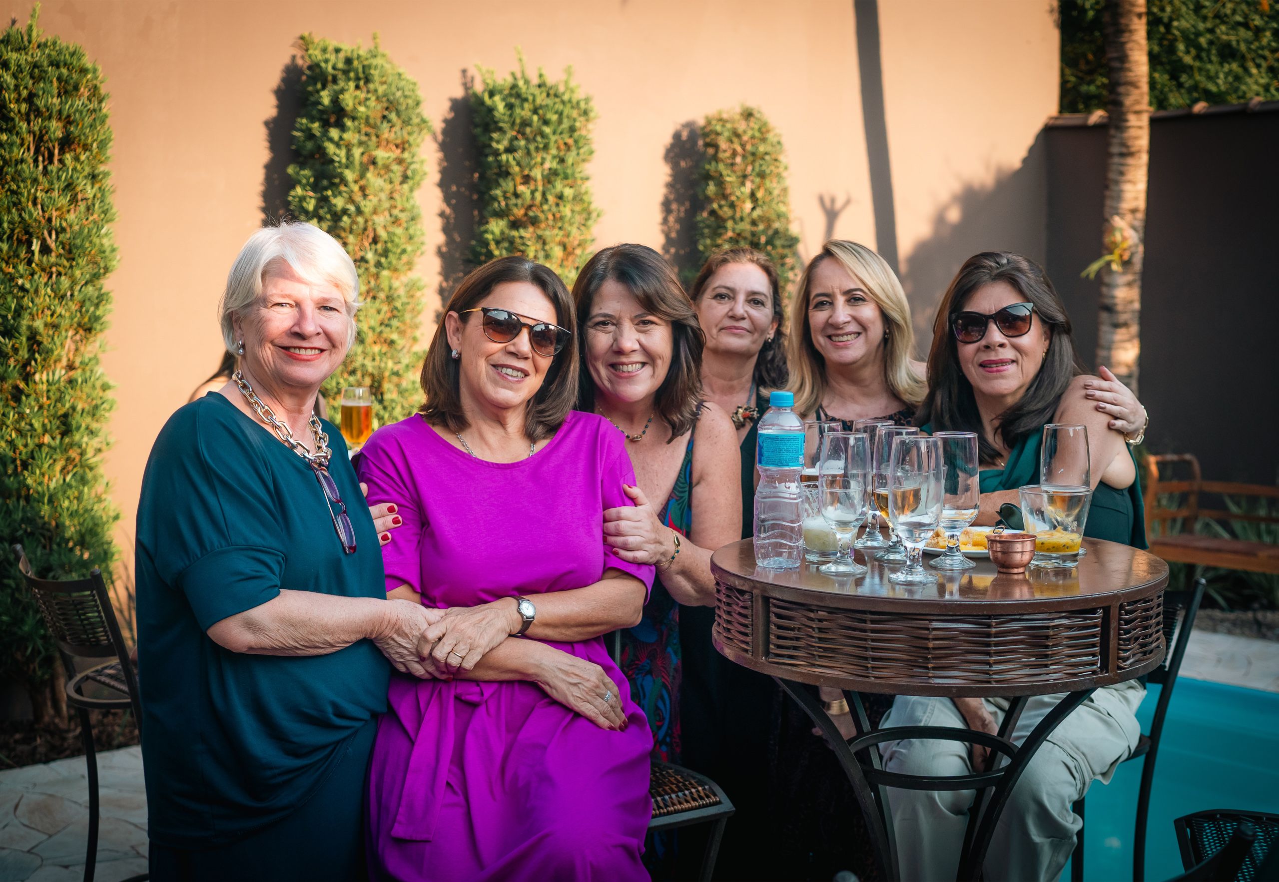 Seis mulheres reunidas ao redor de uma mesa alta com copos, em área externa.