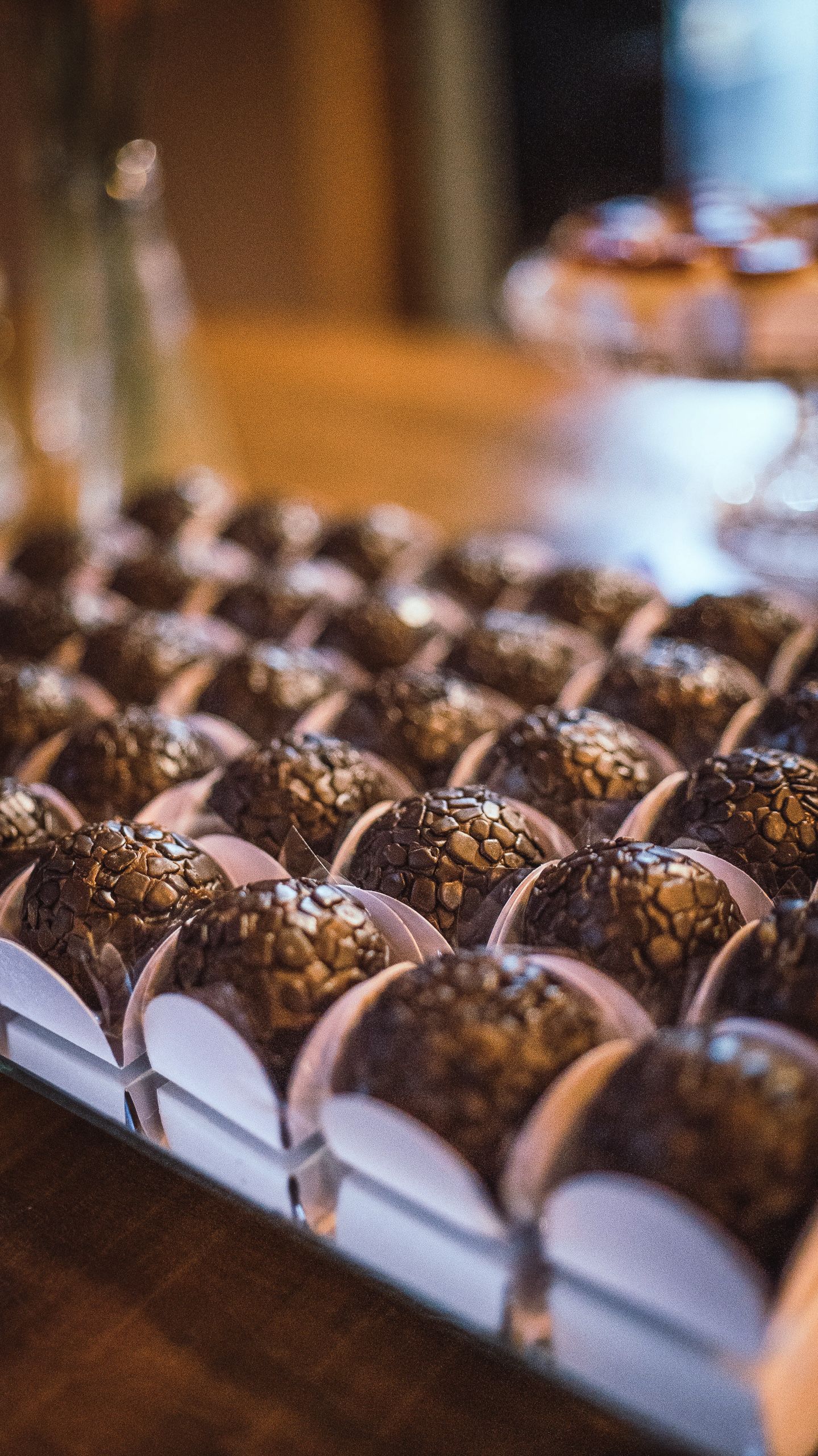 Bandeja de doces de chocolate em forminhas brancas, com mesa de festa desfocada atrás.
