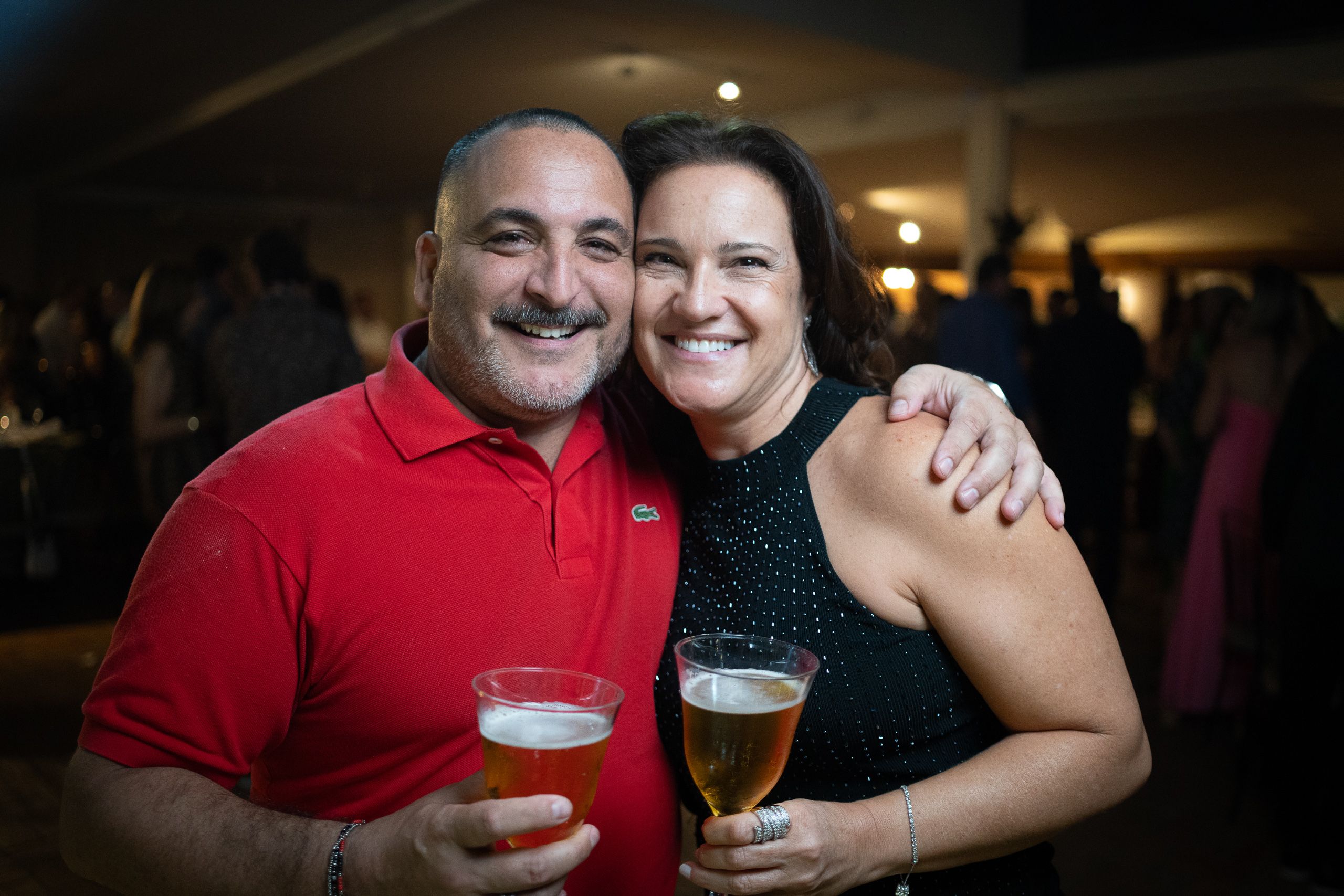 Homem de polo vermelha e mulher de blusa preta brilhante brindando com cerveja em festa.