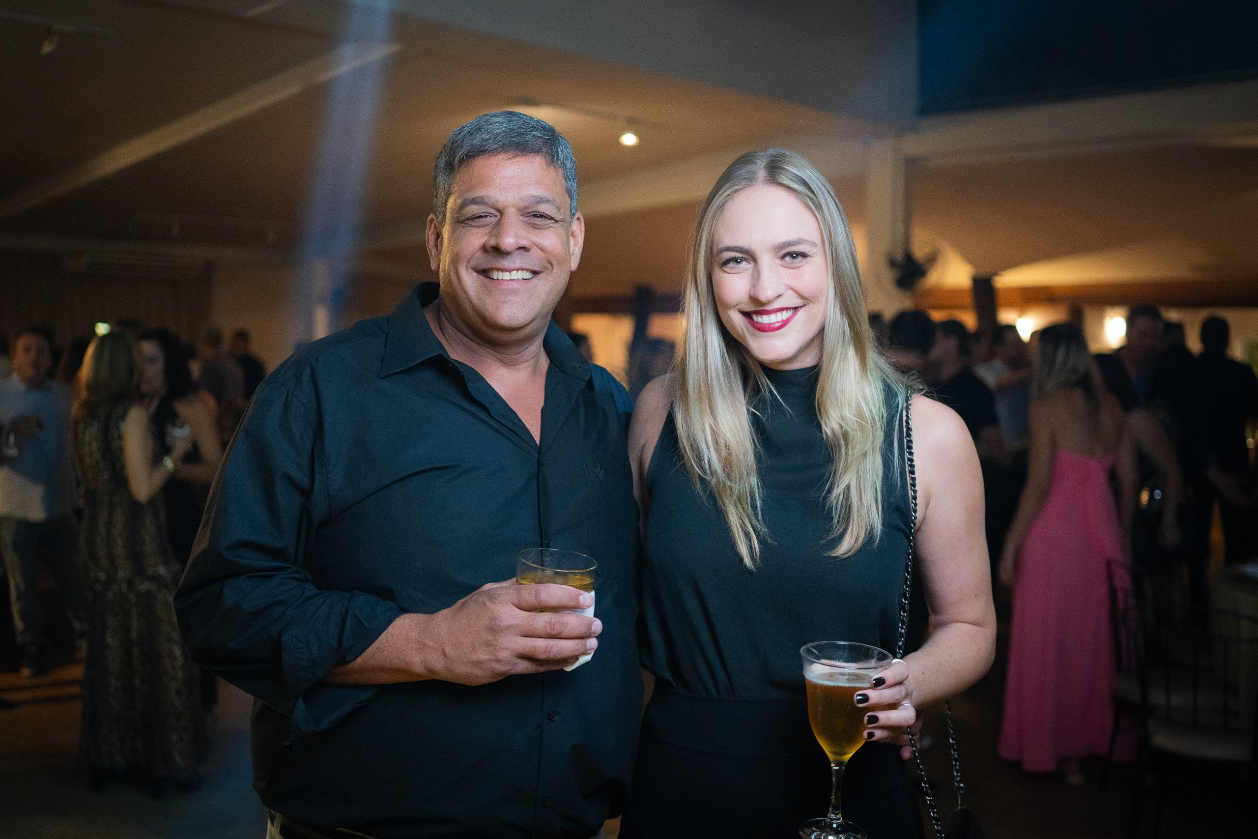 Homem de camisa preta e mulher de vestido preto lado a lado em festa, ambos com bebidas.