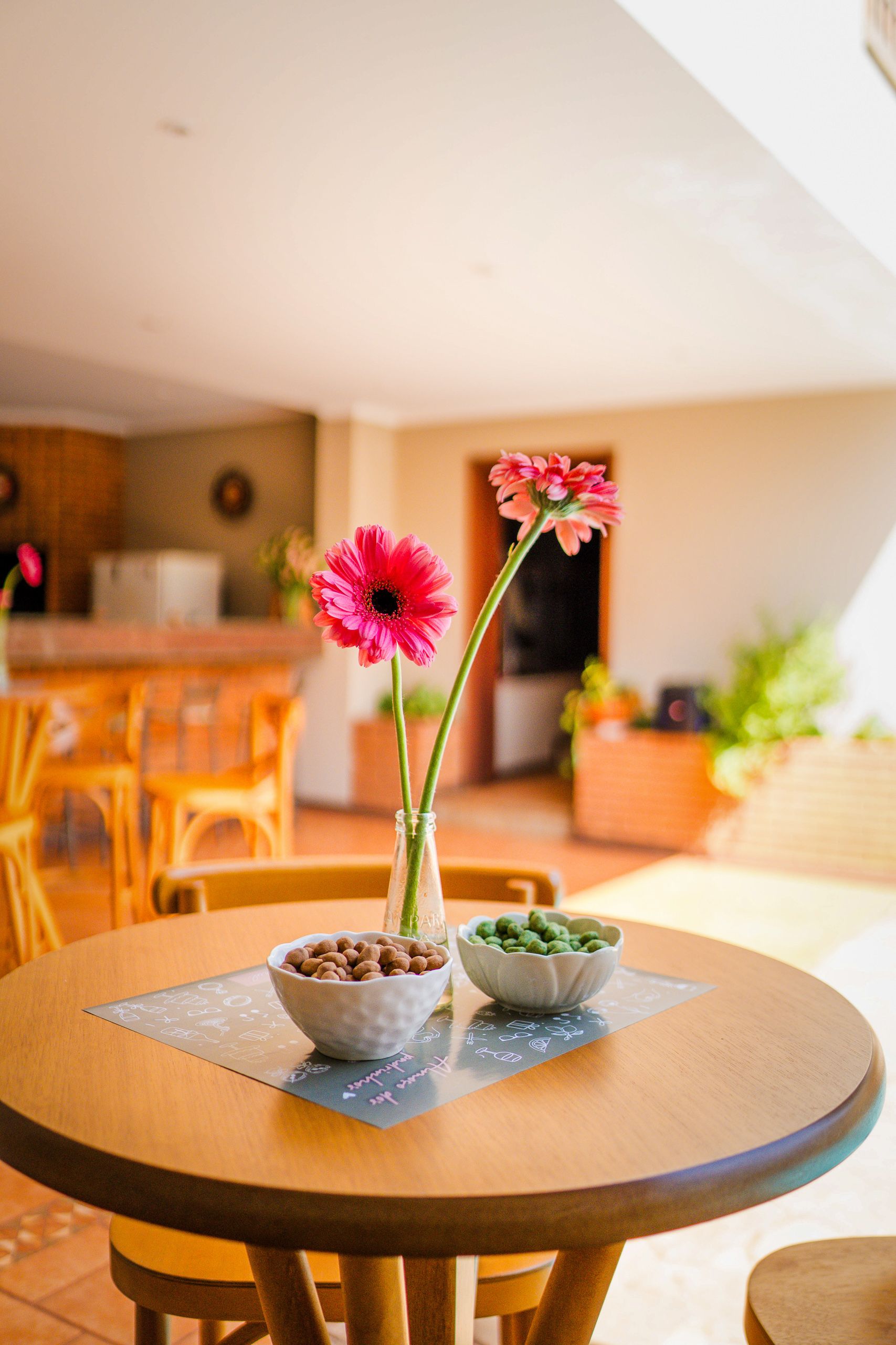 Mesa redonda de madeira com gérberas rosa em garrafa e duas tigelas de petiscos.