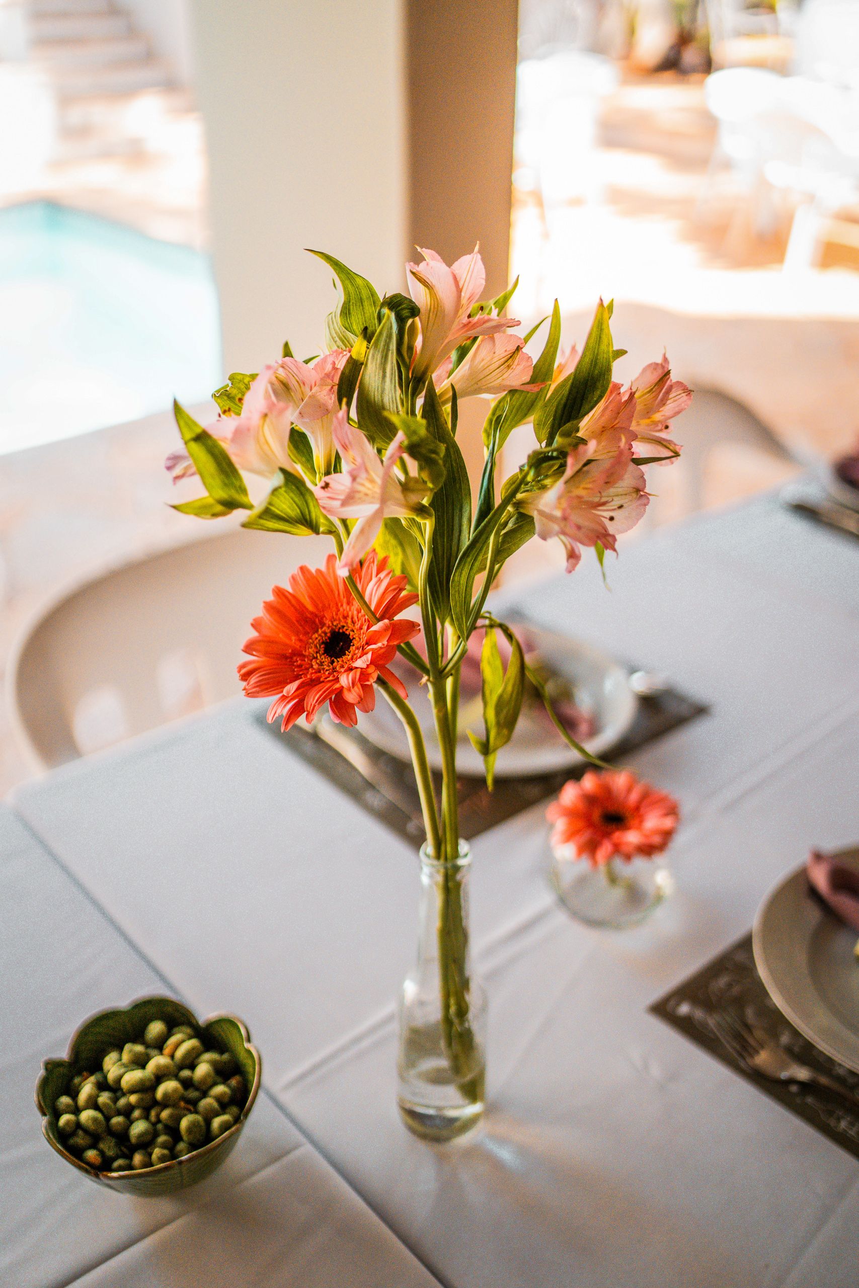 Arranjo de flores rosa e laranja em garrafa de vidro sobre mesa posta, piscina ao fundo.