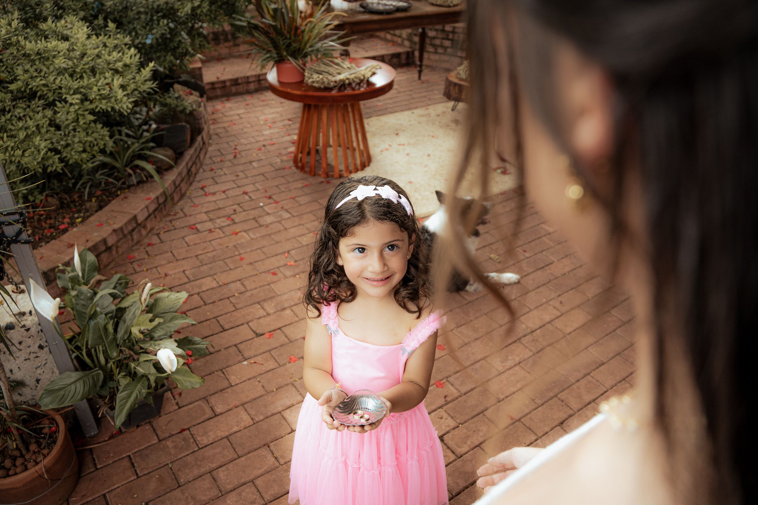Menina de vestido rosa segurando uma concha com alianças, em pátio de tijolos.