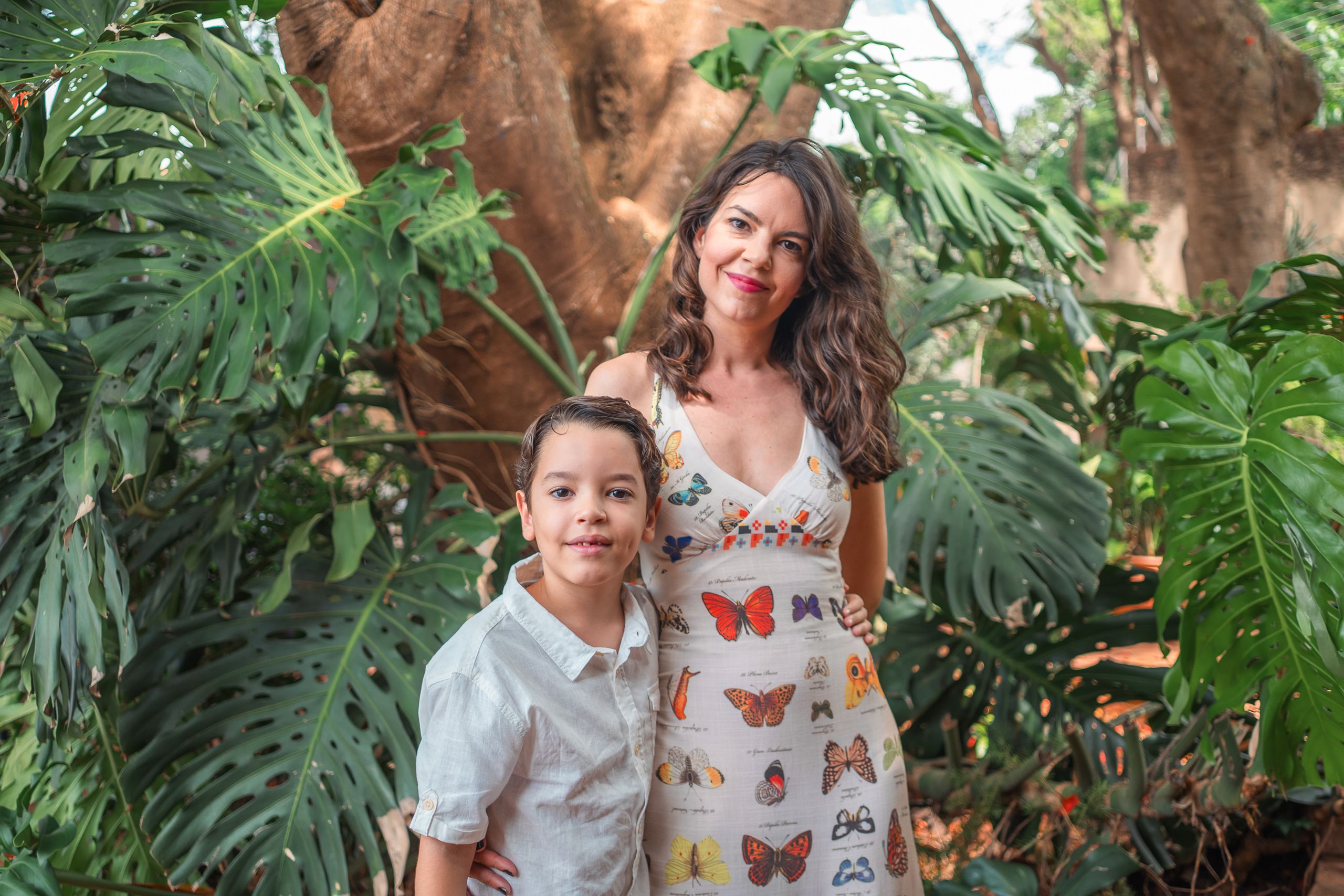 Mulher de vestido estampado e menino de camisa branca entre folhas grandes e tronco de árvore.