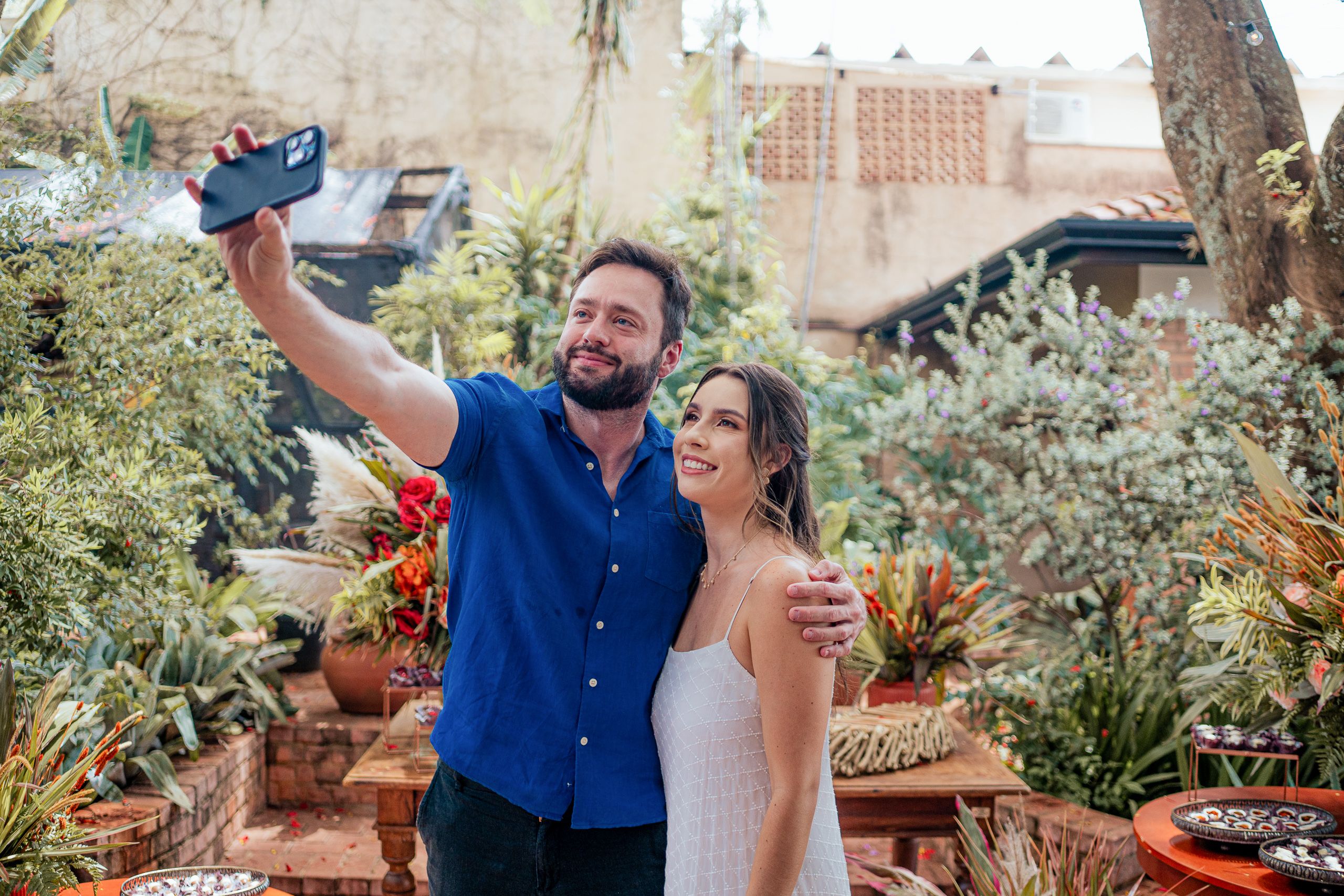 Homem de camisa azul tirando selfie com mulher de vestido branco em jardim decorado.