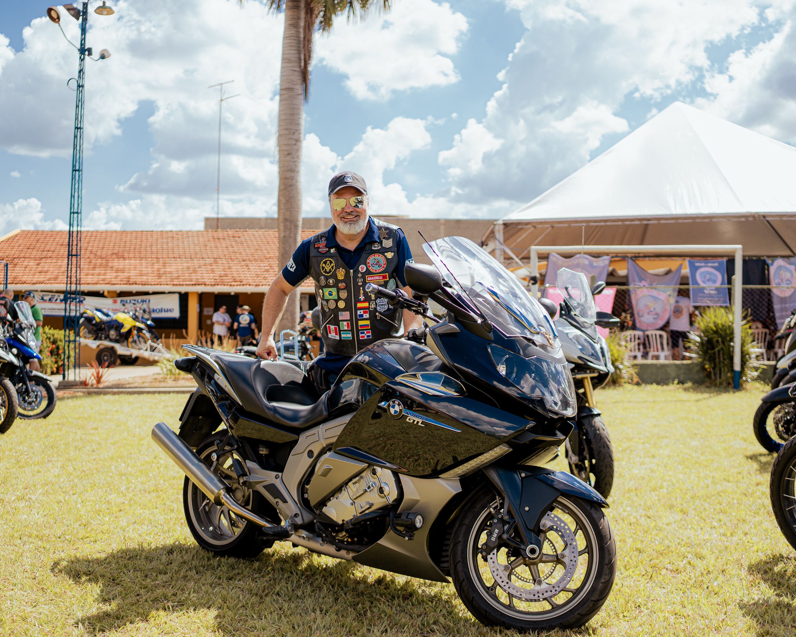 Homem de boné e colete atrás de uma motocicleta grande, em gramado sob céu nublado.