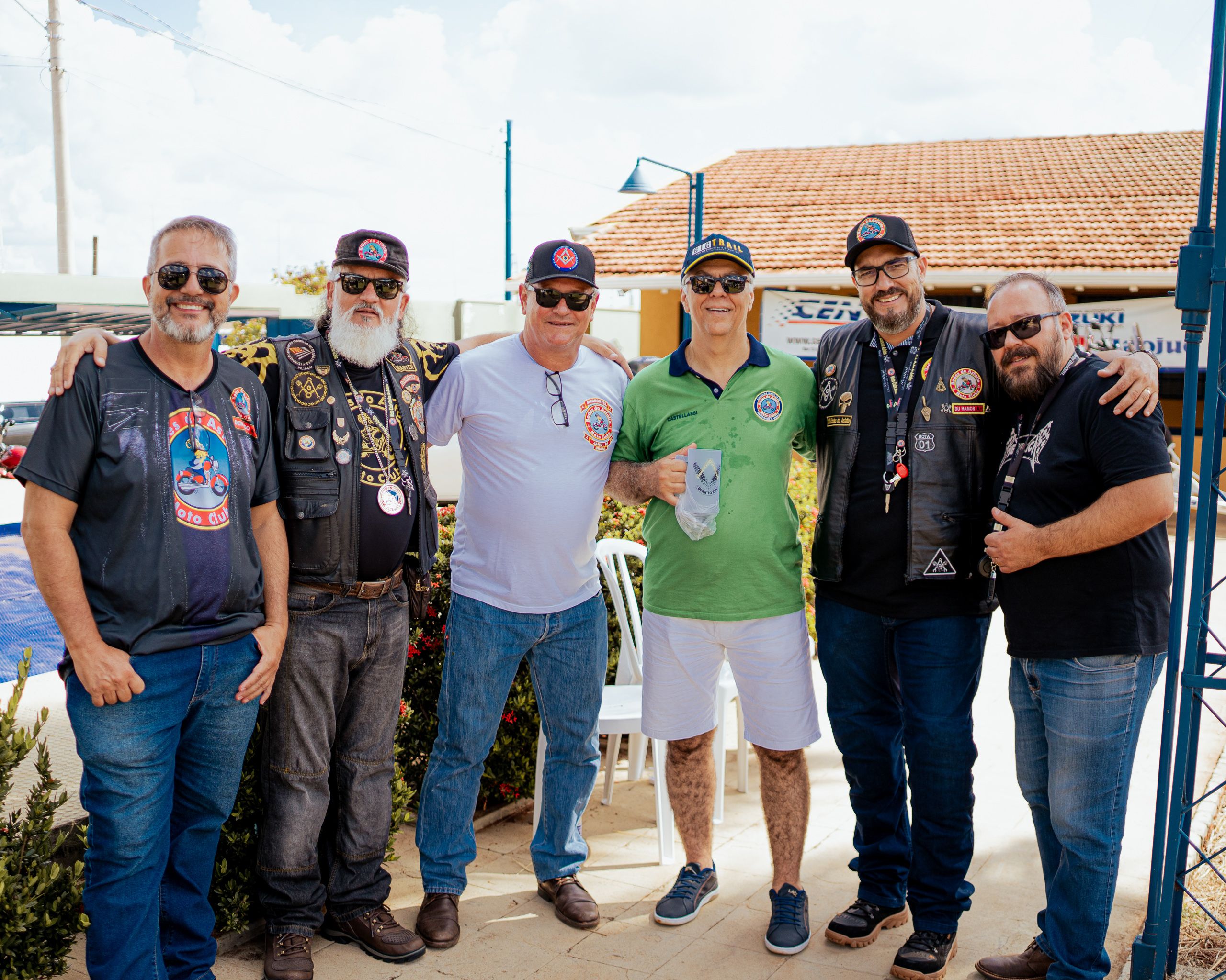 Seis homens de colete e boné posando lado a lado, com telhado e céu ao fundo.