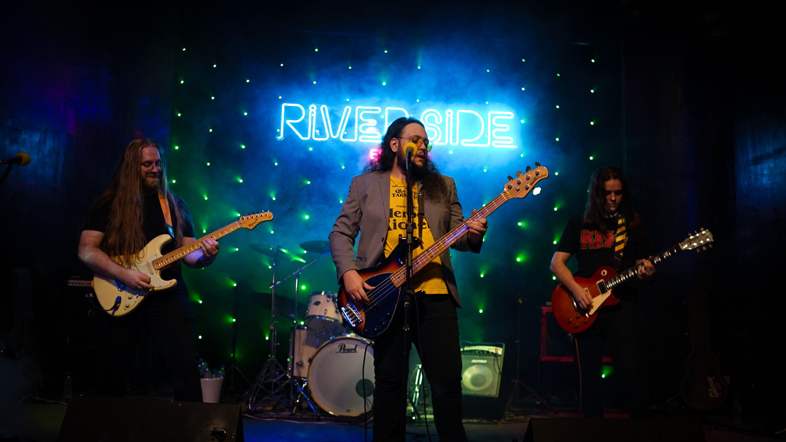 Três músicos tocando em palco sob letreiro de neon azul e luzes verdes.