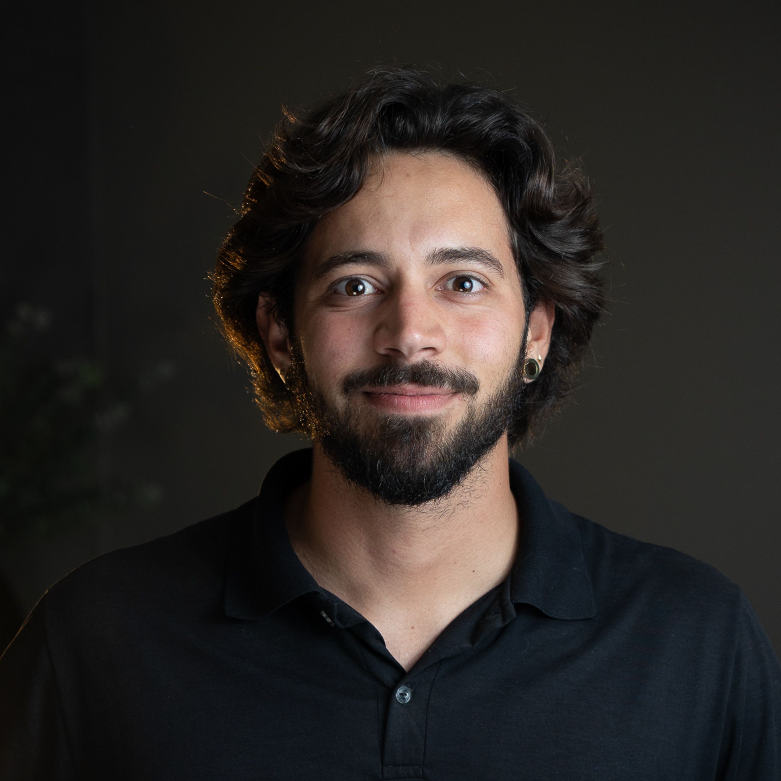 Homem de barba e camisa polo preta, em fundo escuro com luz lateral quente.