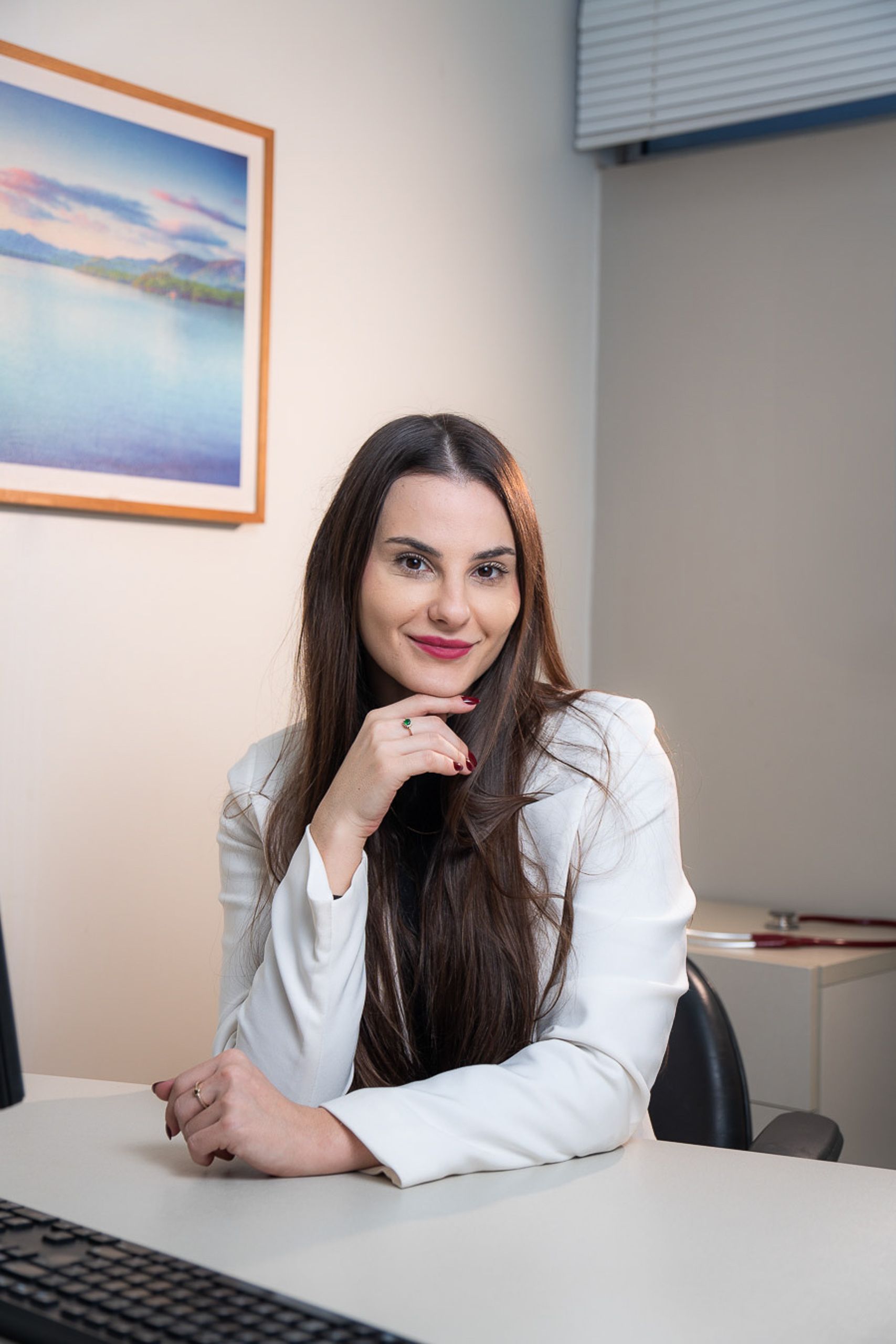 Retrato profissional de médica de blazer branco sentada à mesa, com estetoscópio no móvel ao lado.