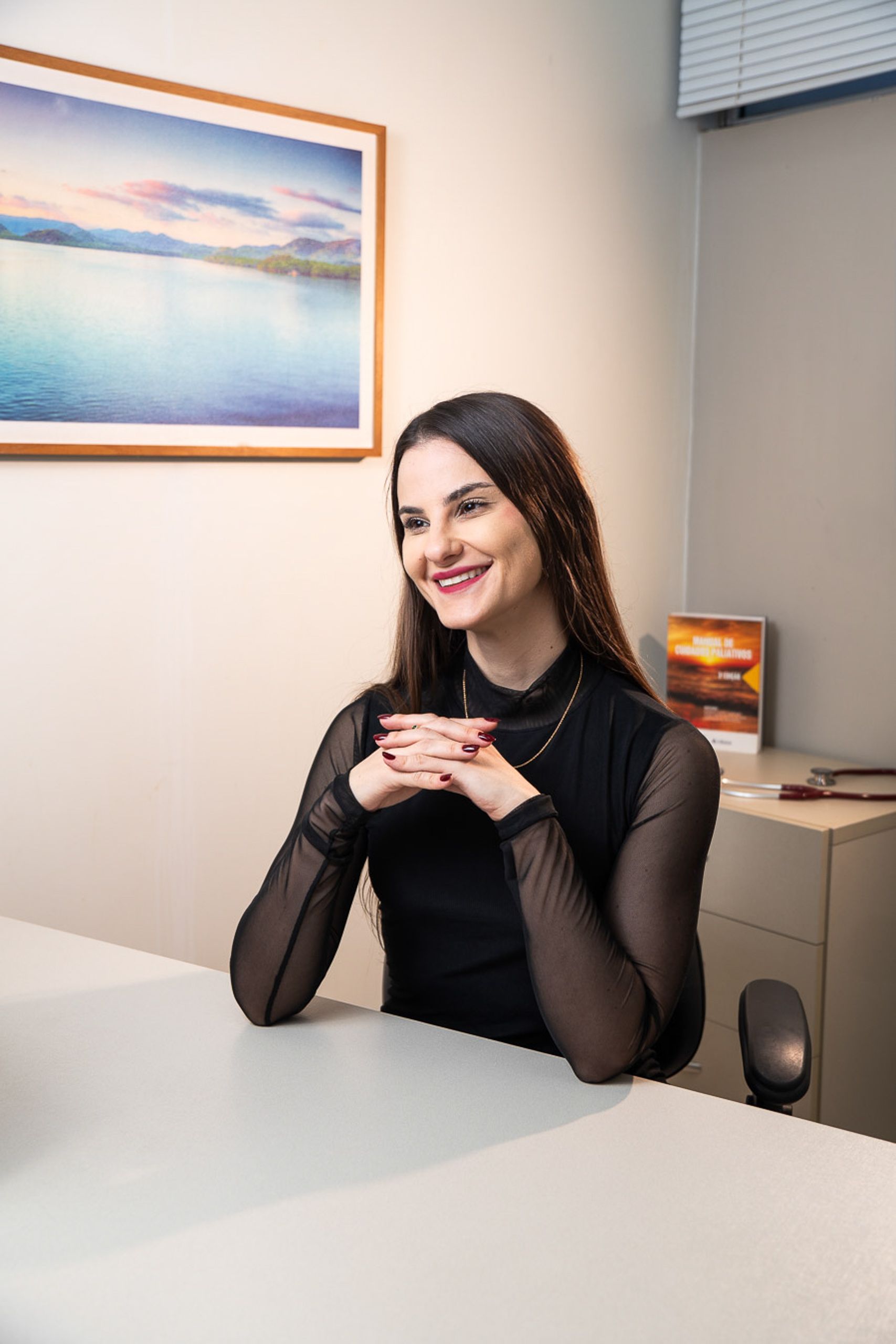 Médica de blusa preta apoiada na mesa do consultório, com livro e estetoscópio ao lado.