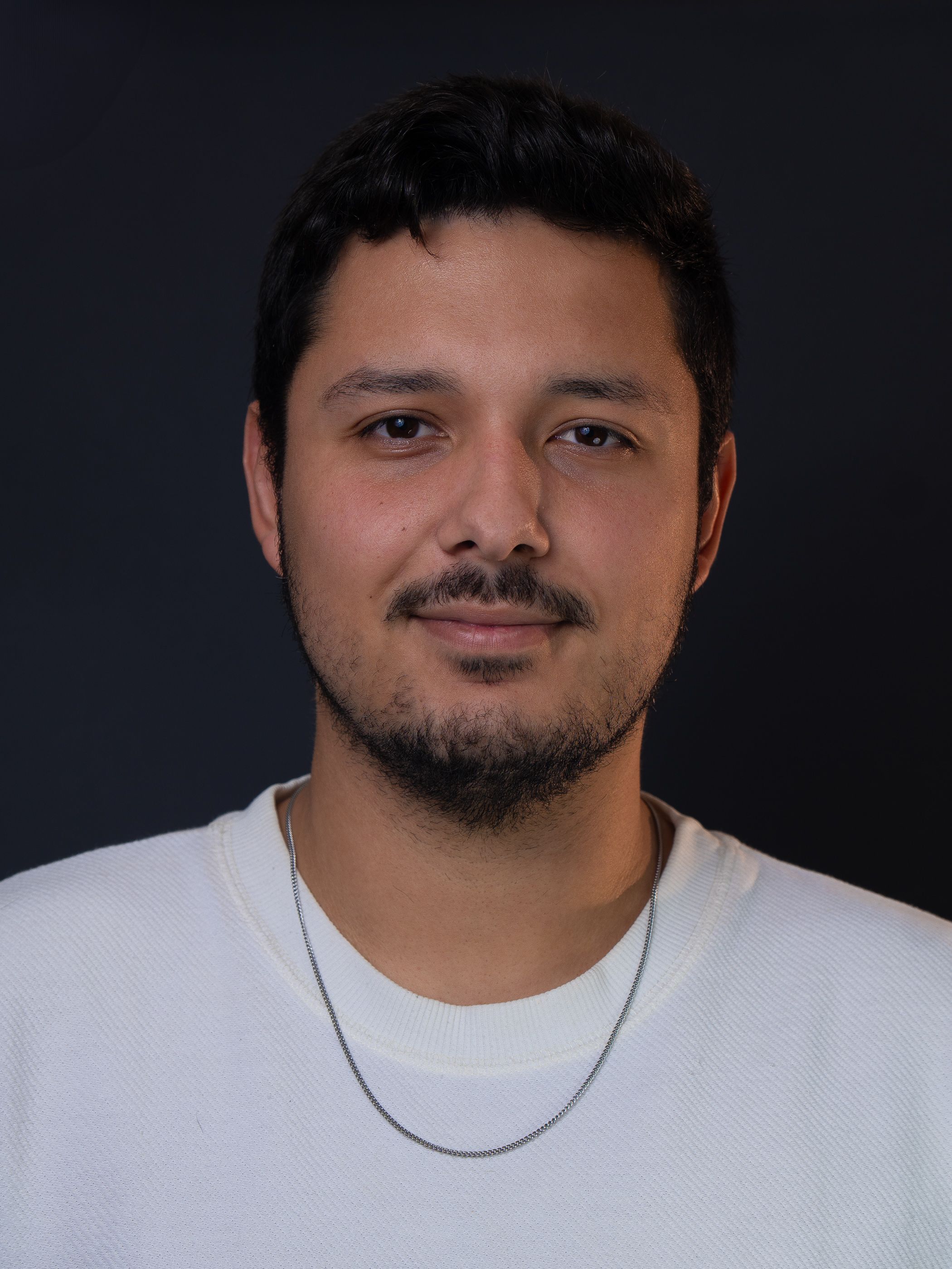 Marcello Naider, de barba e camiseta clara, em retrato de estúdio com fundo escuro.