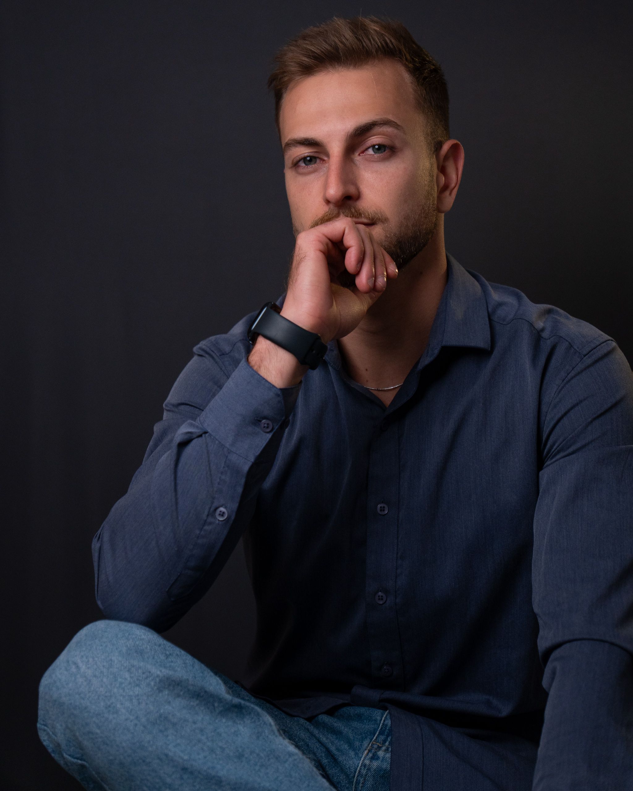 Homem jovem de barba curta sentado com uma perna cruzada, apoiando o queixo na mão fechada, de camisa social azul-marinho, calça jeans e relógio de pulseira preta, em estúdio de fundo cinza-escuro.
